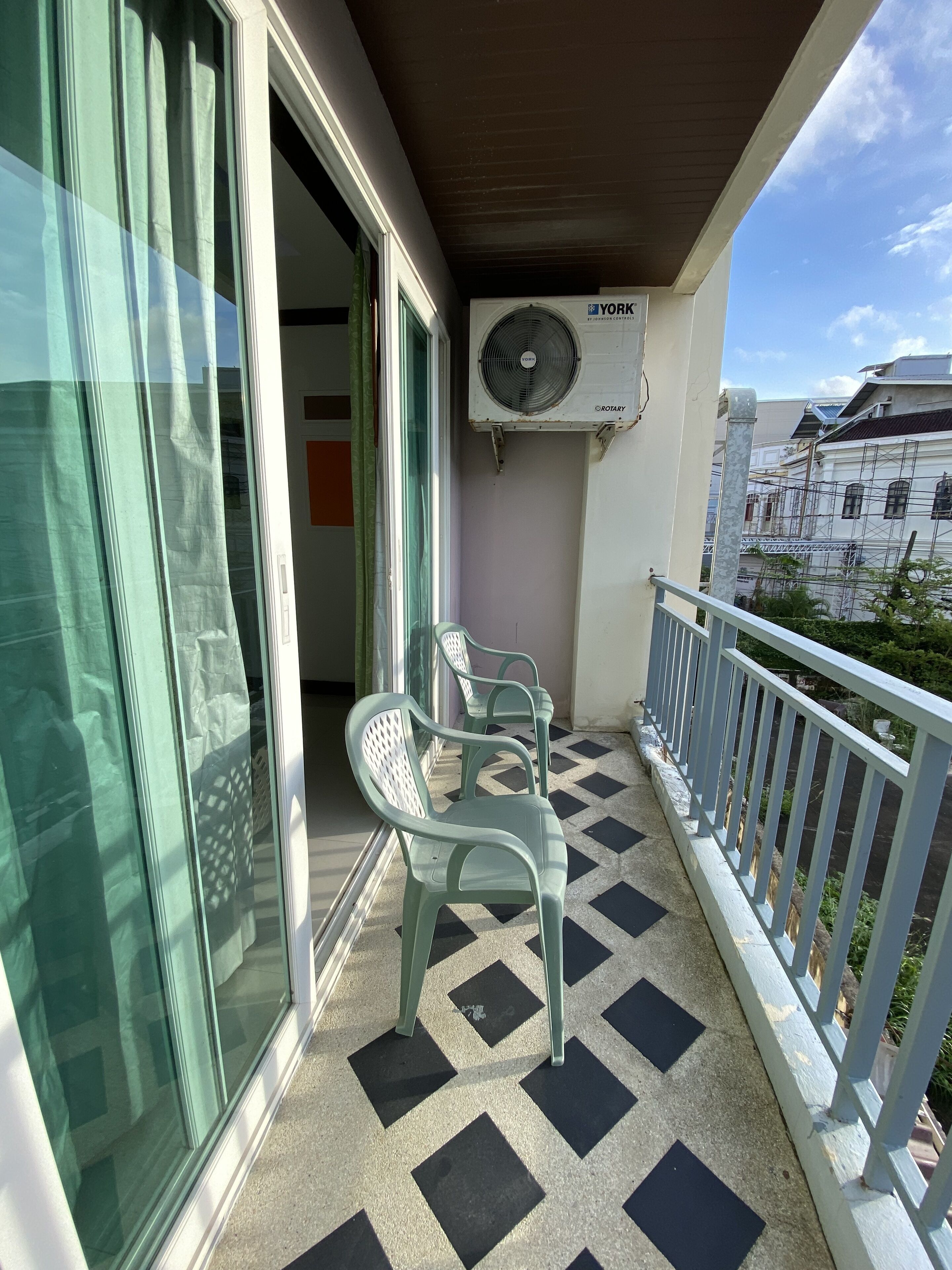 A balcony with a checkered floor, two green plastic chairs, a white railing, and a ceiling fan.