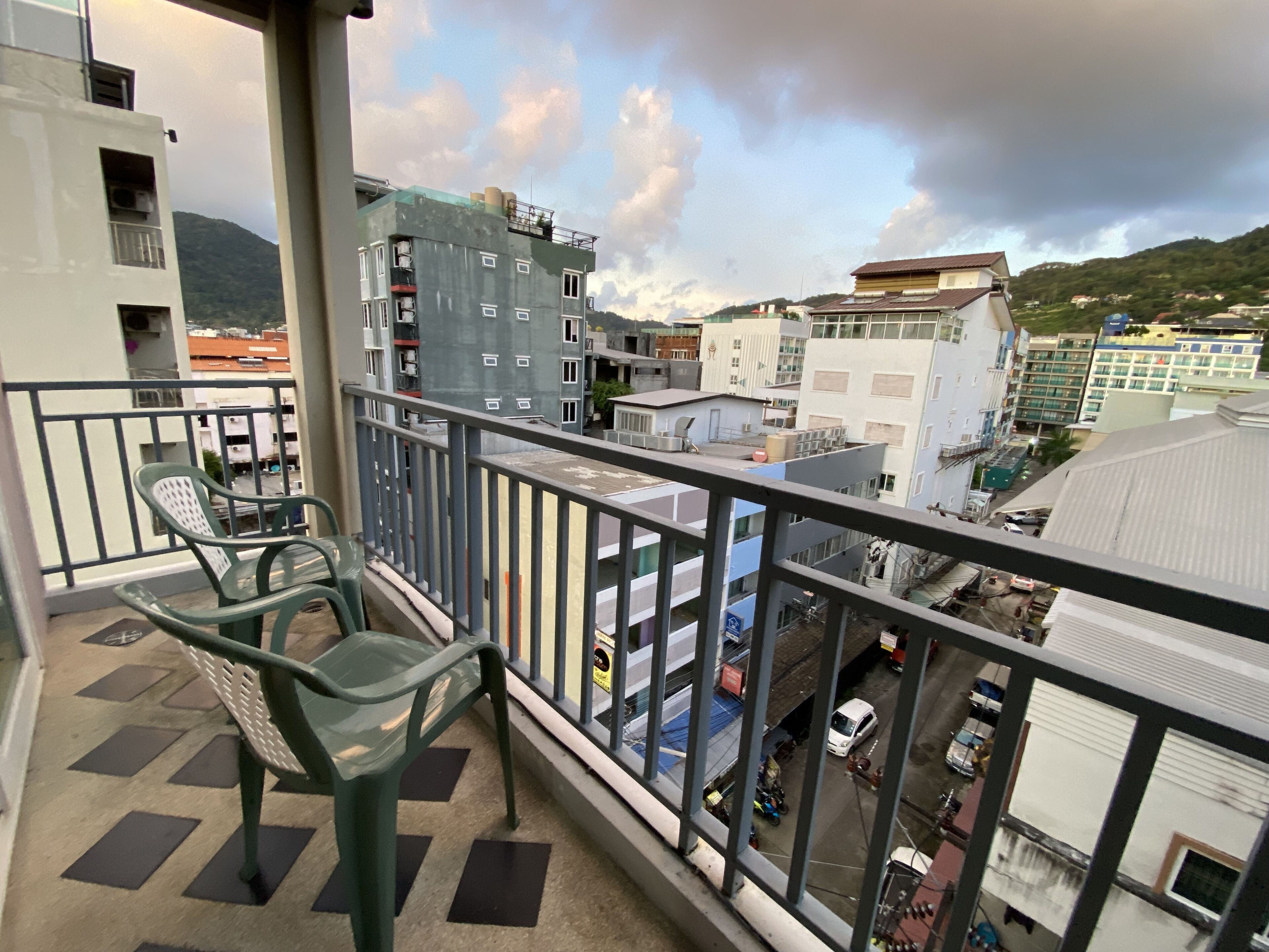 Un balcón con dos sillas de plástico verde, vista de un paisaje urbano con edificios y una montaña al fondo.