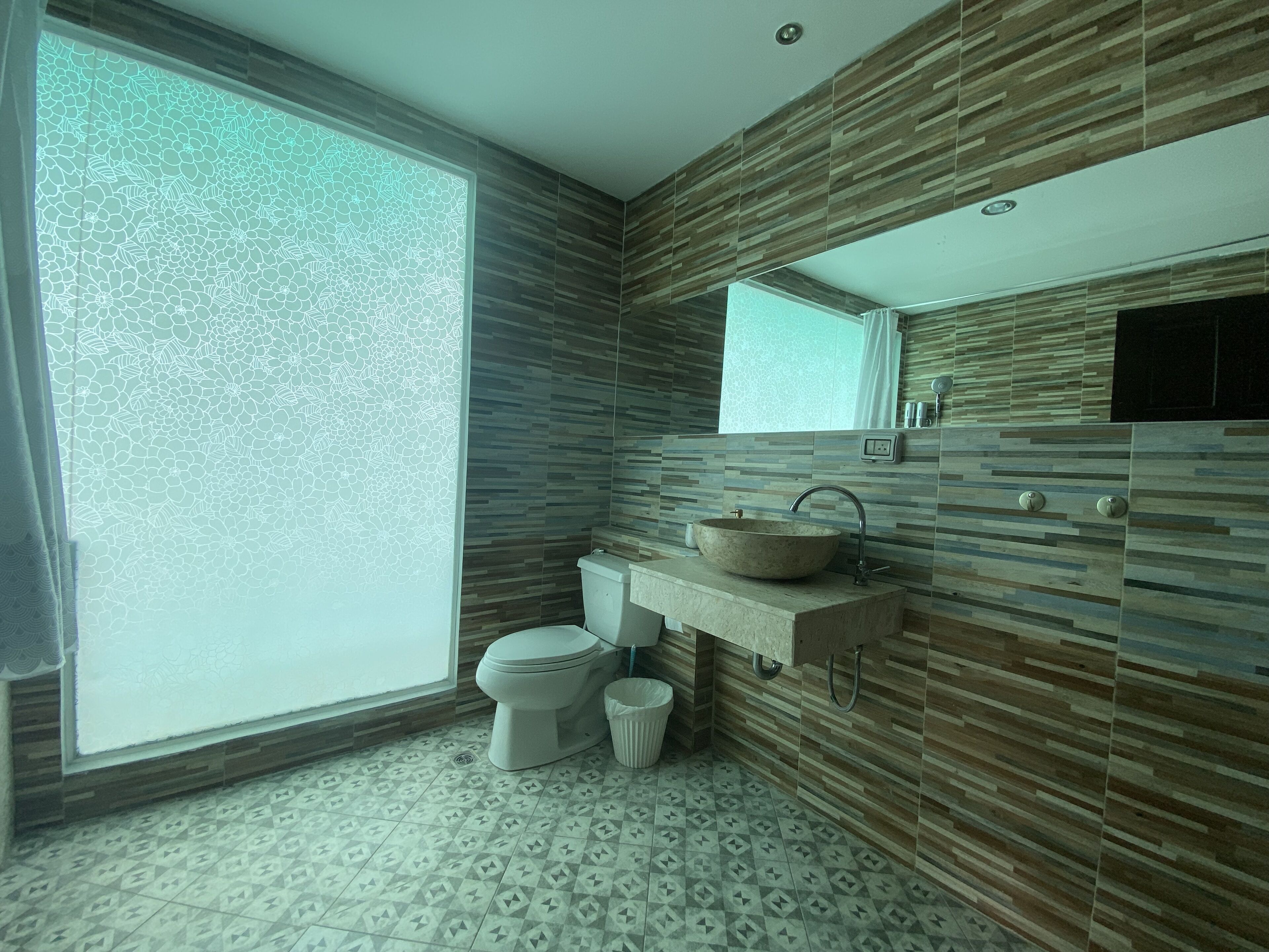 A bathroom with a large window, a sink with a stone basin, a toilet, and a trash bin.