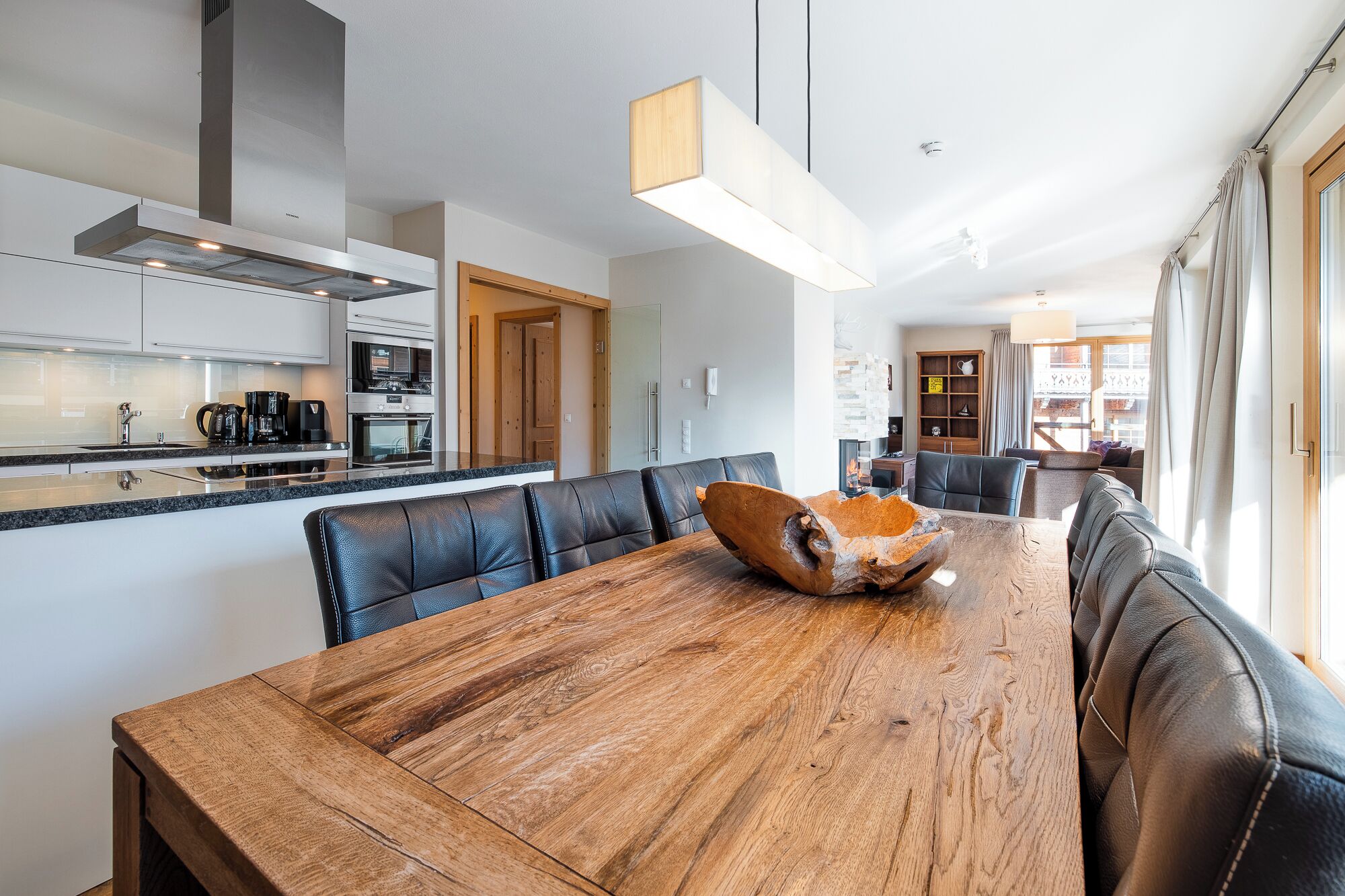 Eine moderne Küche mit Edelstahlgeräten, ein Essbereich mit Holztisch und Ledersesseln sowie ein Blick auf die Stadt durch große Fenster.
