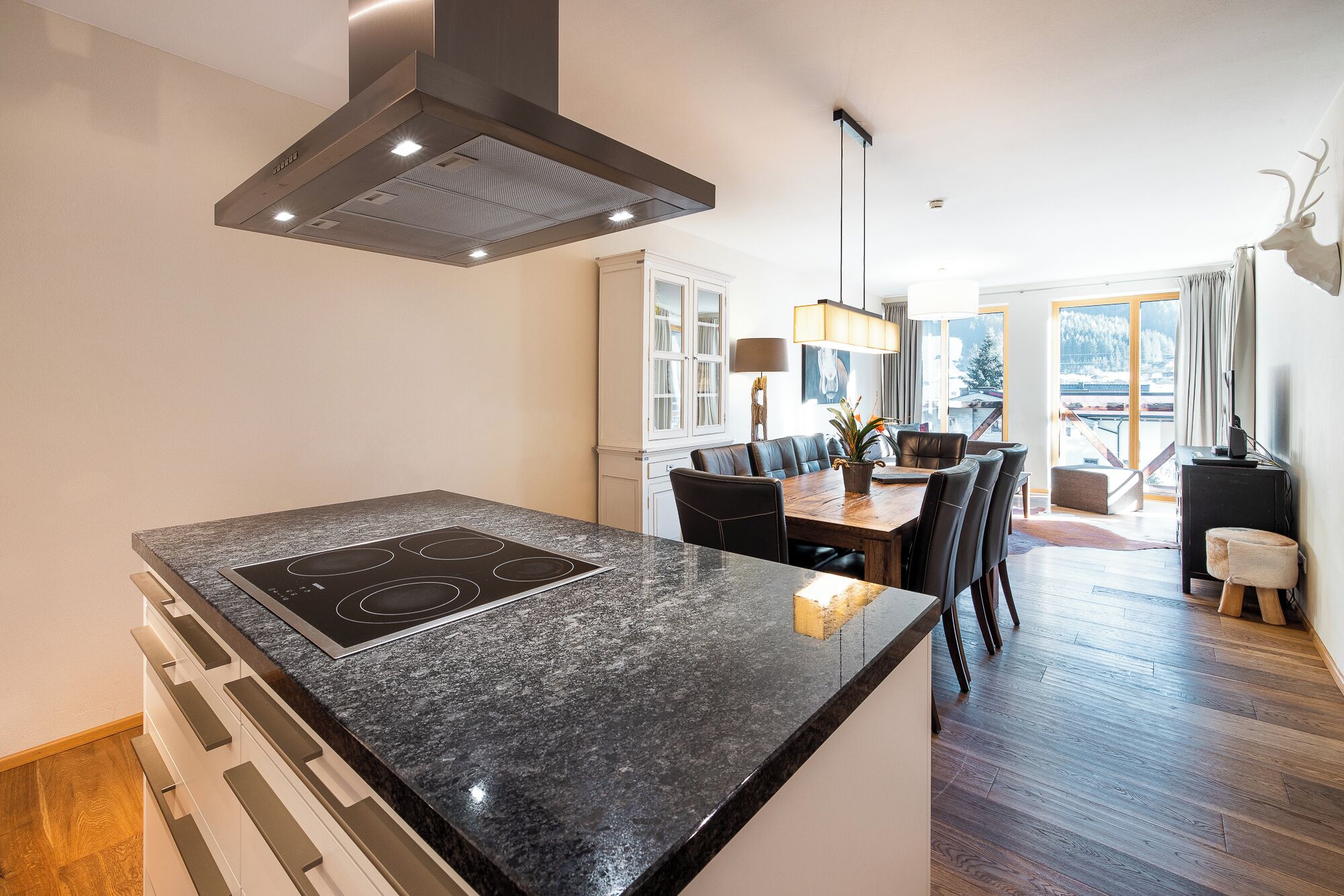 A modern kitchen with a central island, a dining area with a table and chairs, and a view of the outdoors through large windows.