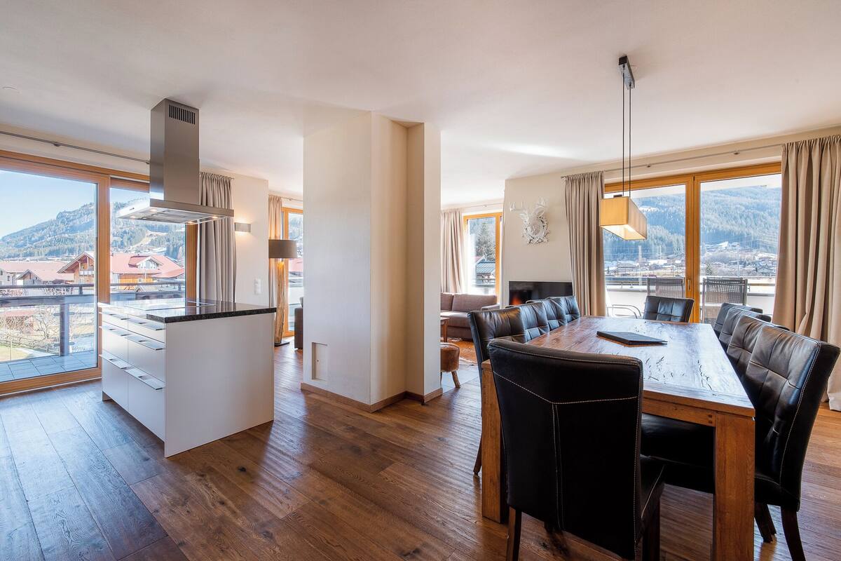 Una cocina moderna con una isla grande, un comedor con mesa de madera y sillas de cuero, y vistas a un paisaje montañoso a través de amplios ventanales.