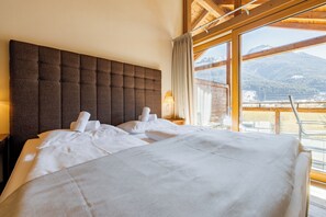 Une chambre d’hôtel avec un grand lit, une tête de lit en bois et une vue sur les montagnes par de grandes fenêtres.