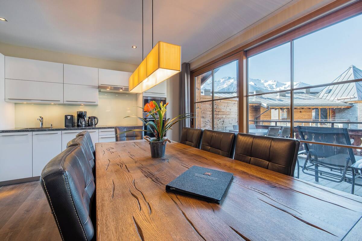 Una cocina moderna con armarios blancos, una mesa de comedor de madera y vistas a las montañas a través de amplios ventanales.