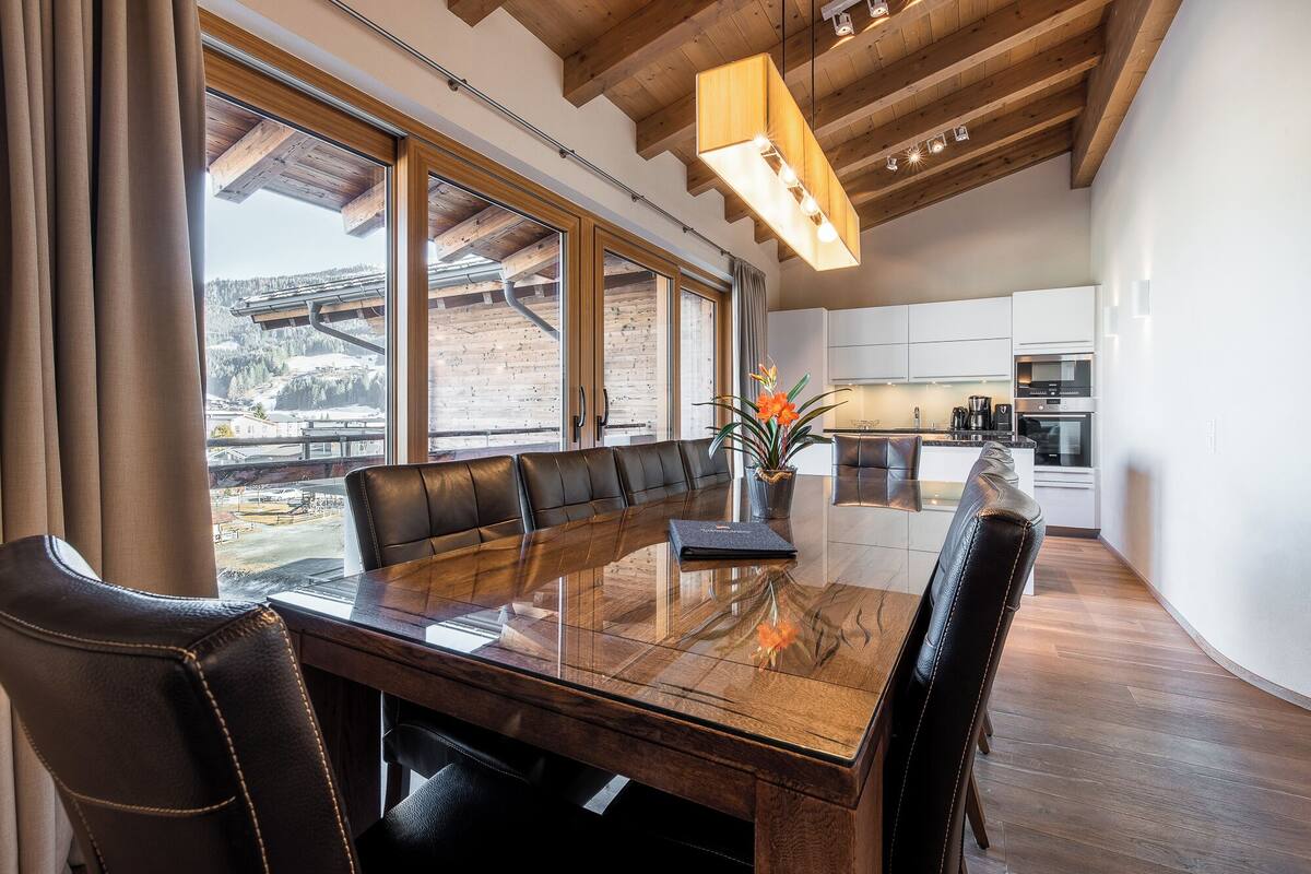 Un comedor con una larga mesa de madera y sillas de cuero, ventanas amplias con vistas a un paisaje nevado y una cocina moderna al fondo.