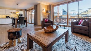Ein geräumiger Wohnbereich mit Couch, Esstisch und Stühlen sowie einem Holztisch. Große Fenster bieten Blick auf eine Berglandschaft.