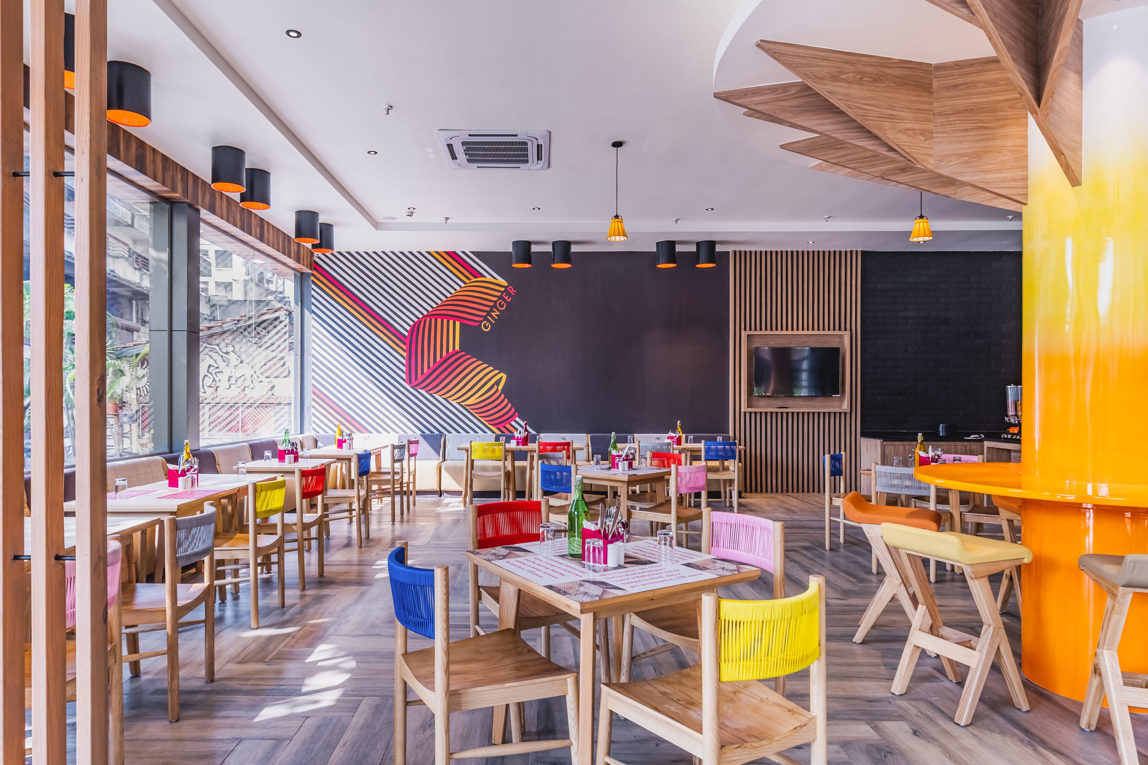Un restaurante moderno con mesas y sillas de madera, sillas coloridas y un diseño de pared vibrante.