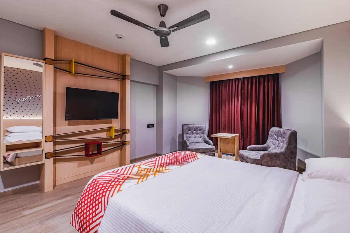A hotel room with a bed, two chairs, a TV, a ceiling fan, a red curtain, and a wooden shelf with towels.