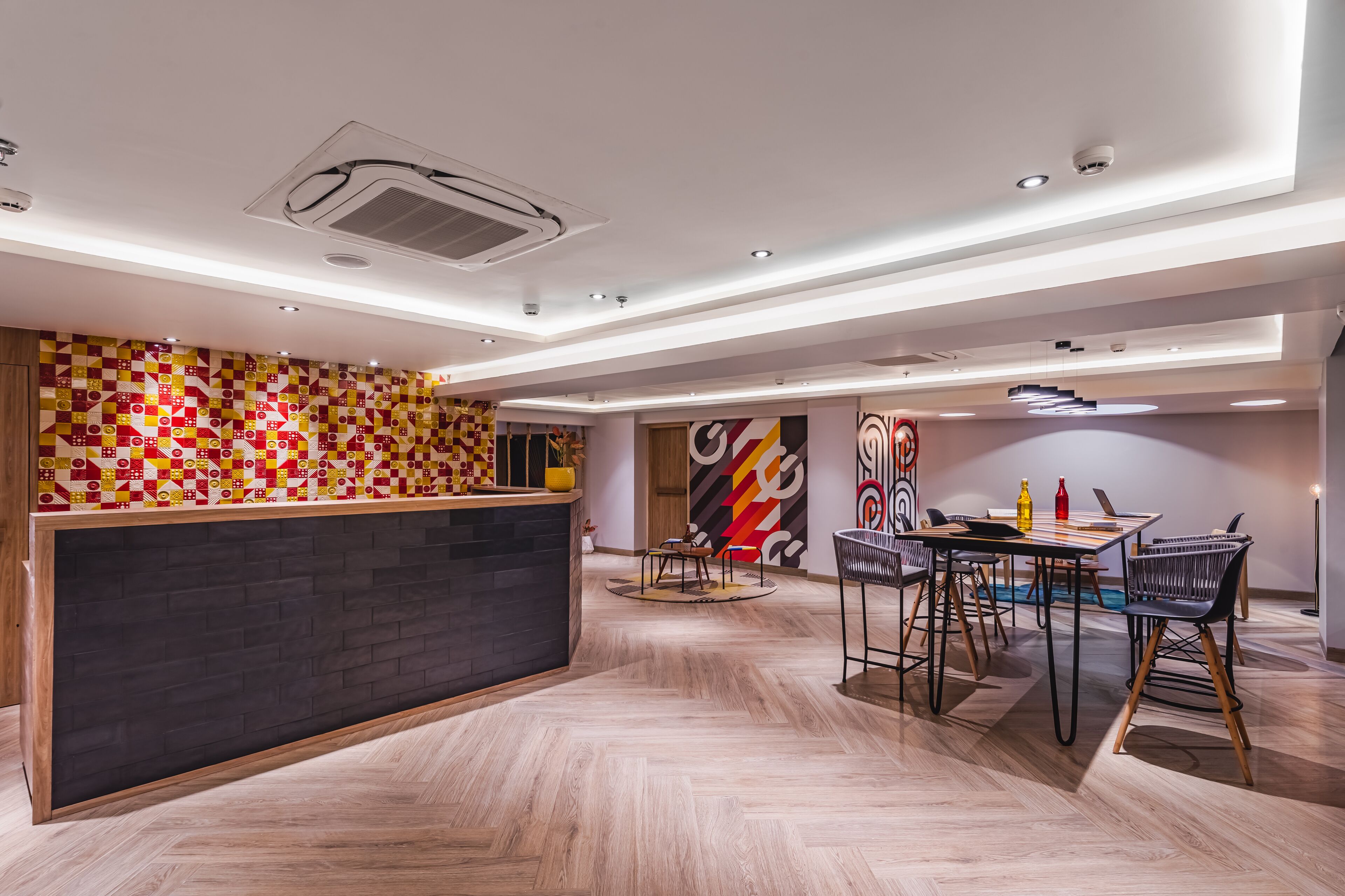 A modern hotel lobby with a reception desk, a dining area with chairs and tables, and a herringbone-patterned floor.