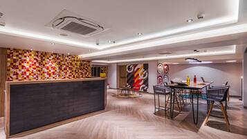 A modern hotel lobby with a reception desk, a dining area with chairs and tables, and a herringbone-patterned floor.