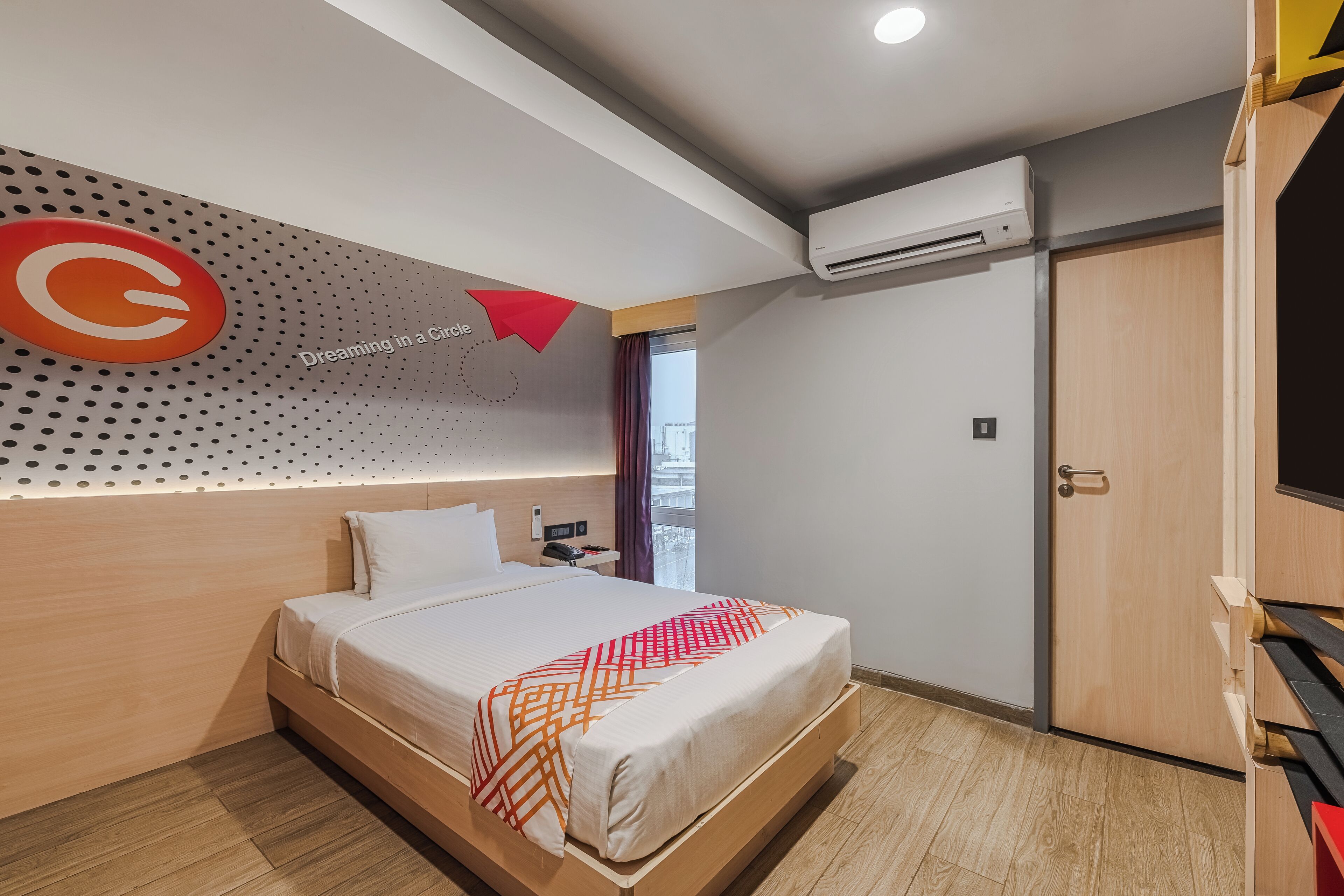 A modern hotel room with a wooden headboard, a large bed with a patterned throw, a wall-mounted air conditioner, and a door with a wooden finish.
