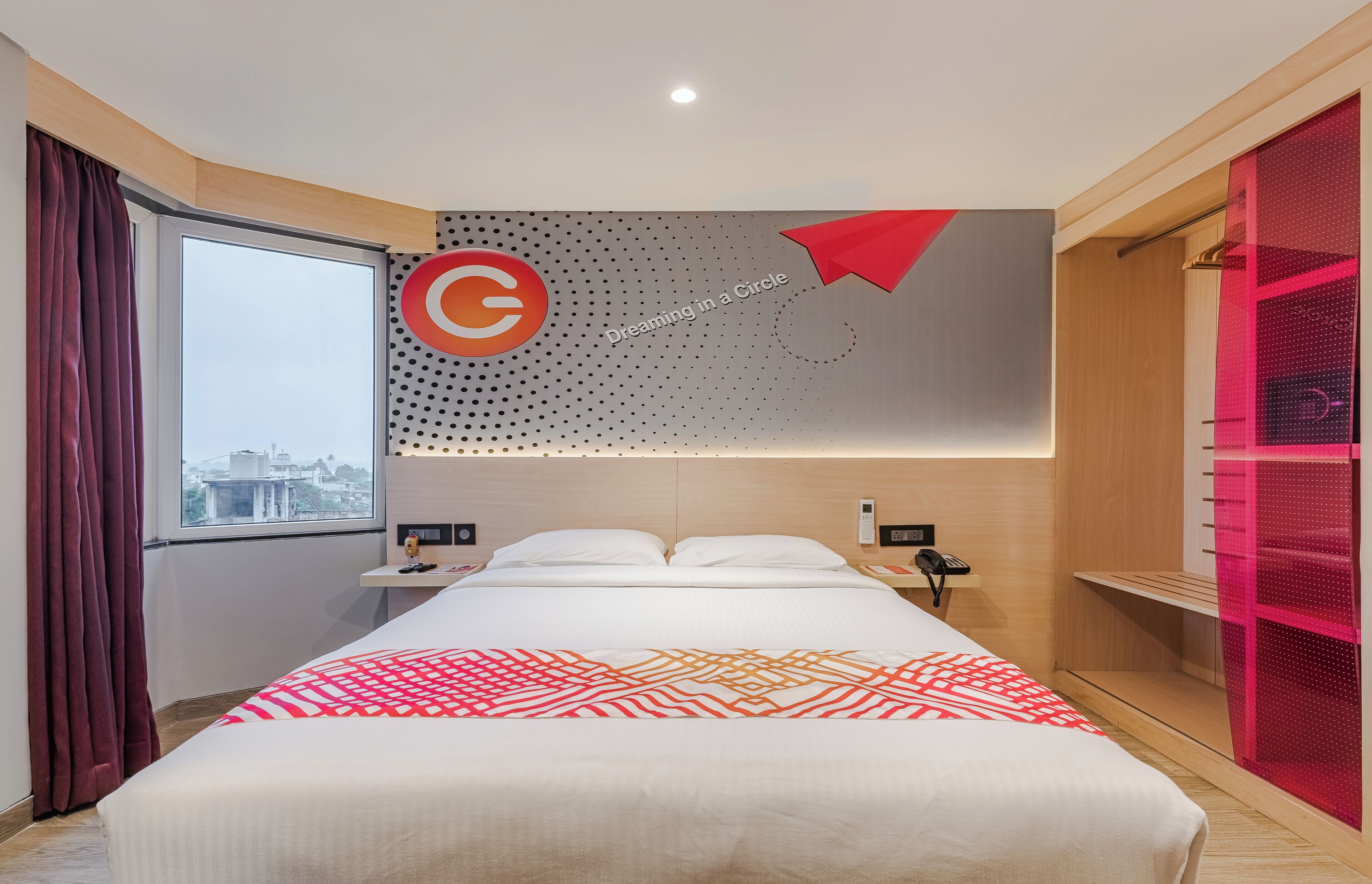 Habitación de hotel moderna con una cama grande, vistas a la ciudad, una colcha de cuadros rojos y blancos, y una pared con un logotipo circular y el texto 'Dreaming in a Circle'.