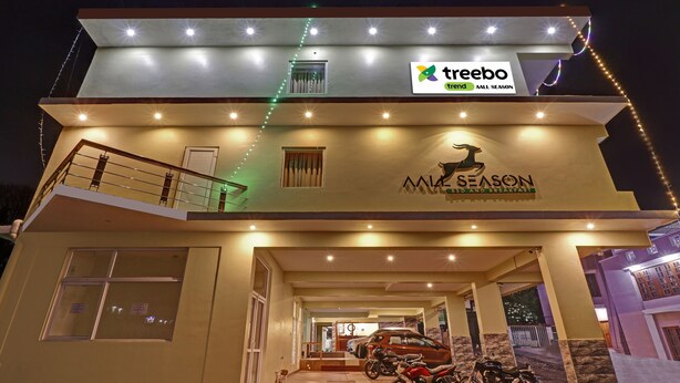 Un hotel con un letrero que indica 'Treebo Trend All Season' y 'M1 Season Bed and Breakfast'. Presenta una estructura de dos pisos con un balcón en el nivel superior, un área de recepción en la planta baja y varios vehículos estacionados.