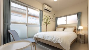 A hotel room with a bed, a chair, a small table, an air conditioner, and a view of the outside balcony.