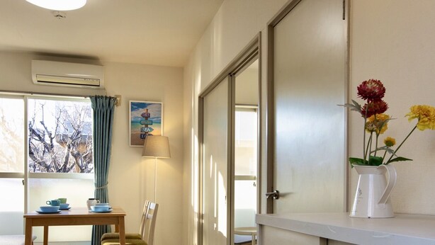 A room with a dining table, chairs, a lamp, a window with curtains, a vase with flowers, and a door.