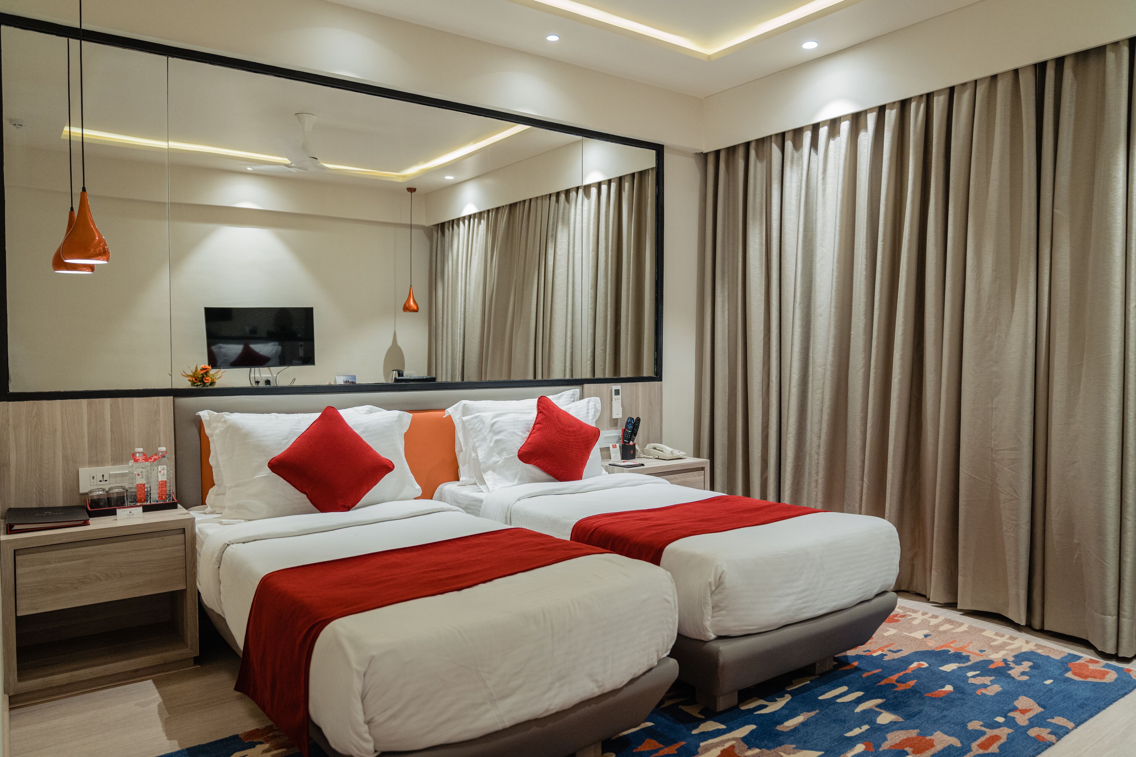 A hotel room with two beds, a large mirror, a flat-screen TV, a bedside table with a lamp, and a window with curtains.