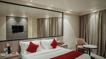 Habitación de hotel con una cama grande, una silla, una mesita, una mesita de noche, un espejo y cortinas.