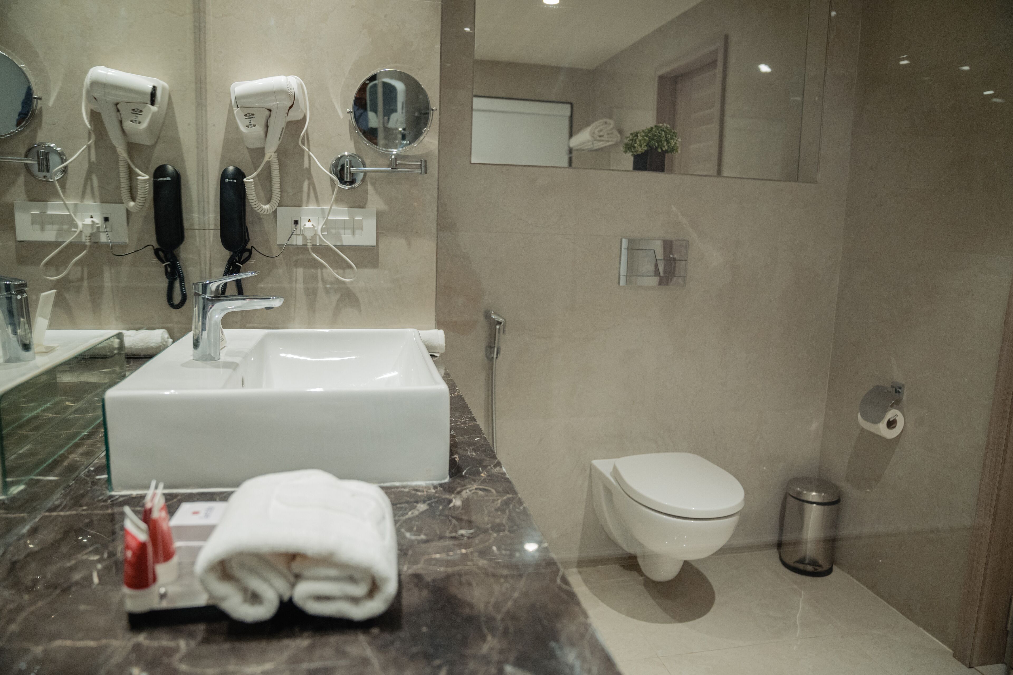 A modern bathroom with a large sink, two hair dryers, a toilet, and a trash can.