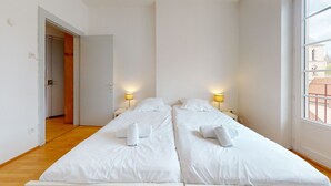 Ein Zimmer mit zwei Betten, Holzboden, einer Tür und einem Fenster mit Blick auf Gebäude.