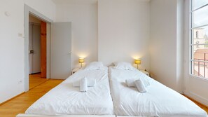 Una habitación con dos camas, suelo de madera, una puerta y una ventana con vistas a edificios.