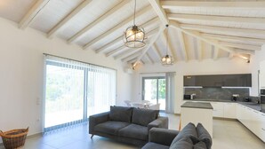 A spacious living area with a modern kitchen, large windows, and a high ceiling with exposed beams. - Villa Loki in Kremithas (Corfu)