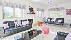 A modern living room with two sofas, a coffee table, and a pink ottoman. There are colorful chairs, a small table, and a baby stroller. The room has large windows and a ceiling light fixture. - Villa Georgios Rafaela (Goudi)
