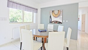 A modern dining room with a round table and six white chairs, a large window with a view of greenery, and two abstract paintings on the wall. - Villa Georgios Rafaela (Goudi)