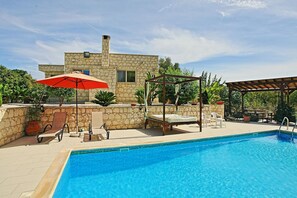 A swimming pool with a stone house in the background, a wooden pergola, and outdoor seating. - Villa Georgios Rafaela (Goudi)