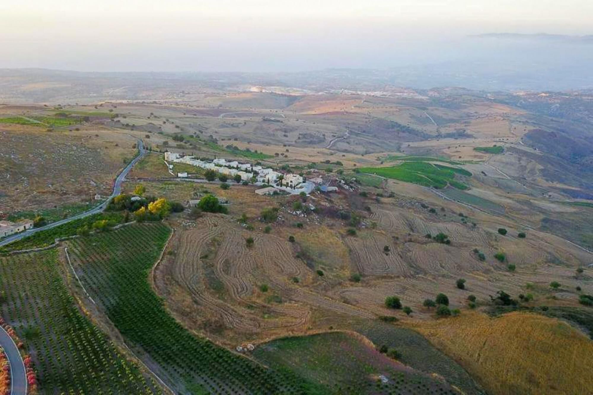 An aerial view of a rural landscape with terraced fields, a winding road, and a cluster of white buildings.
