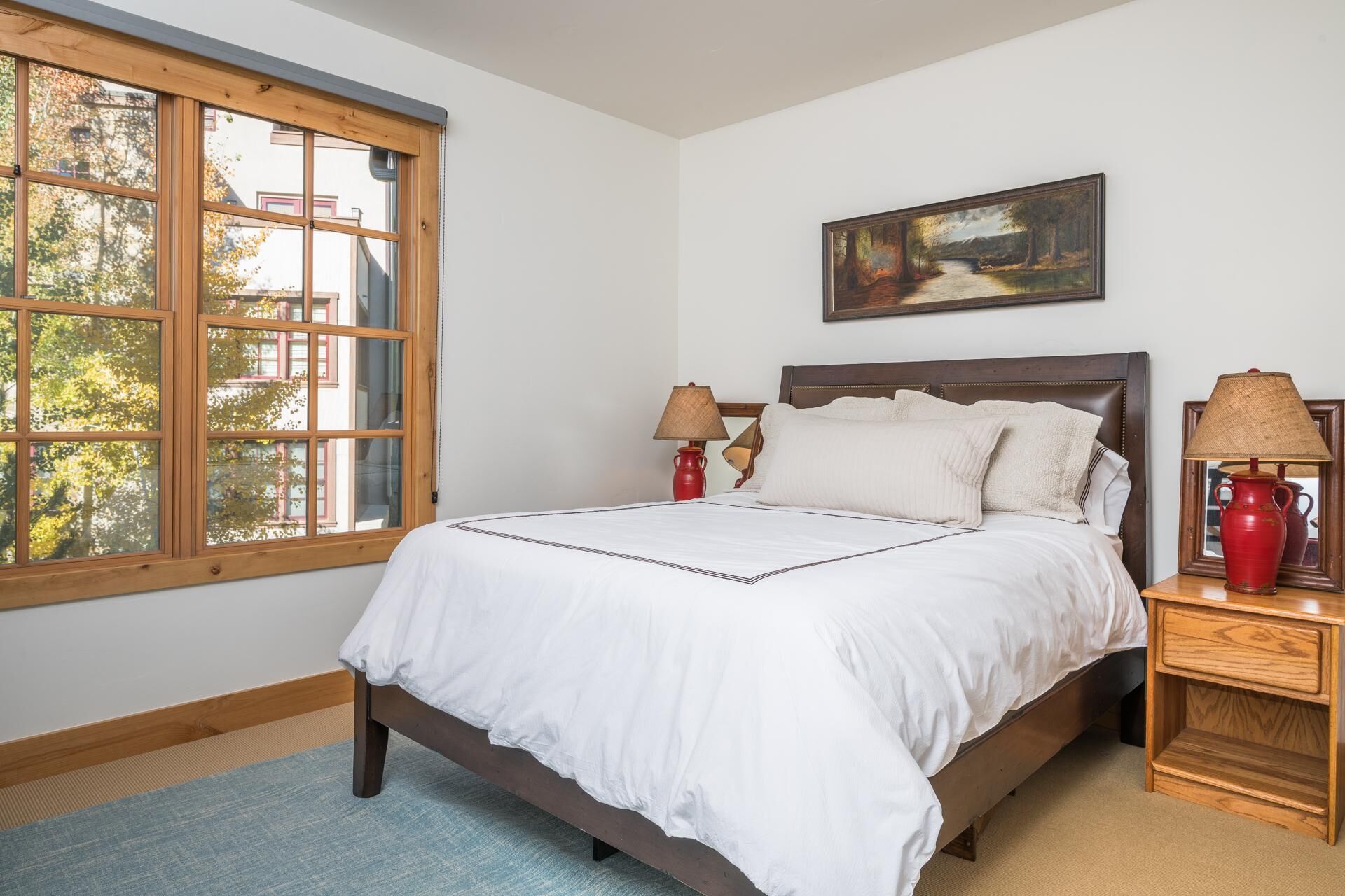 A neatly made bed with white linens, a painting above it, and a window with a view of trees and buildings.