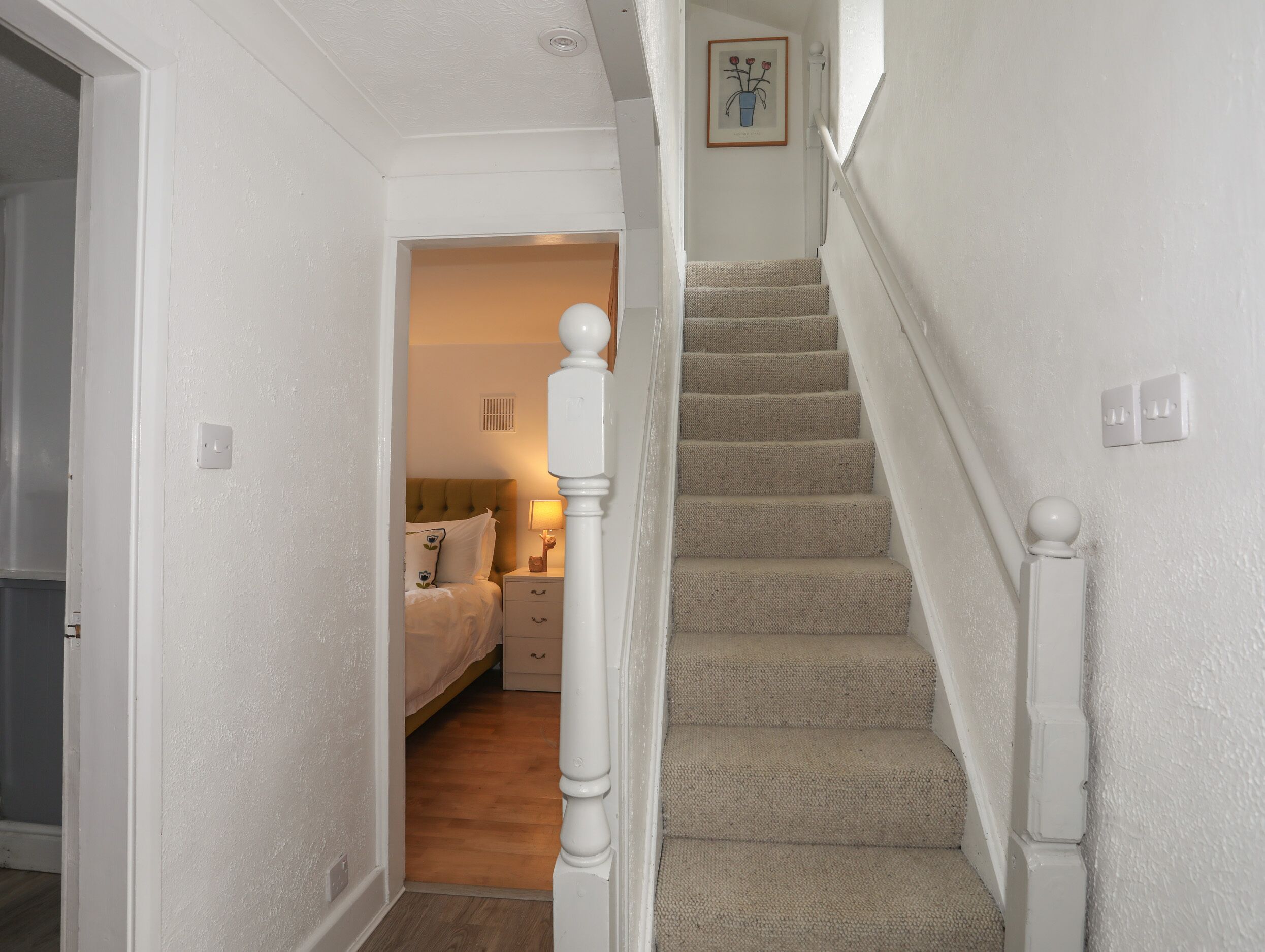 A staircase with a carpeted landing and a bedroom with a bed, nightstand, and lamp.