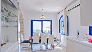 A modern kitchen with white cabinets, a dining area with a wooden table and white chairs, a large window with blue frames, and a hanging light fixture. - Villa Mandola Rosa in Poli Crysochous (Polis)