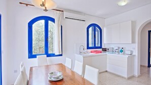 A modern kitchen with white cabinets, a wooden dining table, and two large windows with blue frames. - Villa Mandola Rosa in Poli Crysochous (Polis)