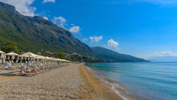 A pebble beach with beach umbrellas and lounge chairs, a clear blue sea, and mountains in the background. - Tinas Apartment in Mparmpati (Corfu)