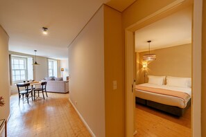 Ein Schlafzimmer mit Bett, Nachttischlampe und Blick auf einen Wohnbereich mit Sofa und Esstisch.