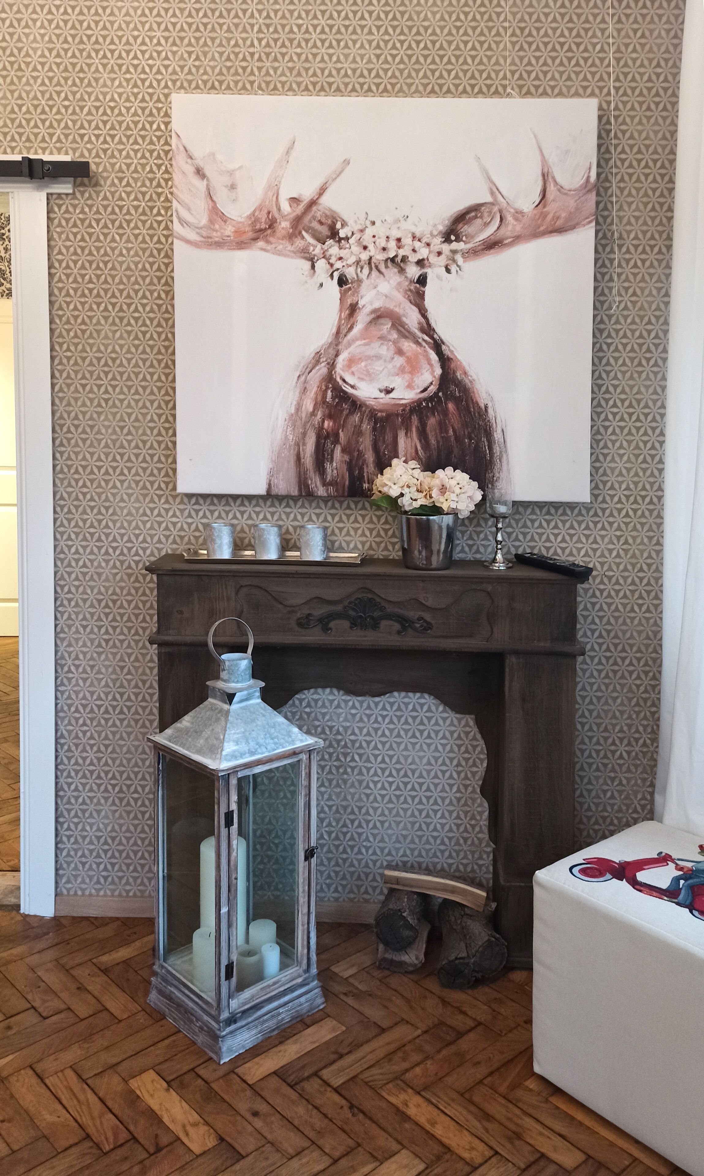 A room with a fireplace, a painting of a moose, a metal lantern, and a table with a red and blue design.