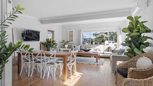 A spacious living room with a wooden dining table, white chairs, a sofa, and a large plant.