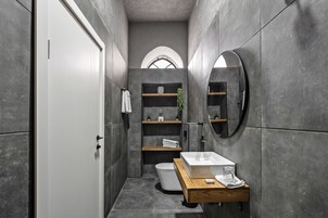 Un baño moderno con un inodoro blanco, un tocador de madera, un espejo y estantes empotrados con toallas y artículos de aseo.