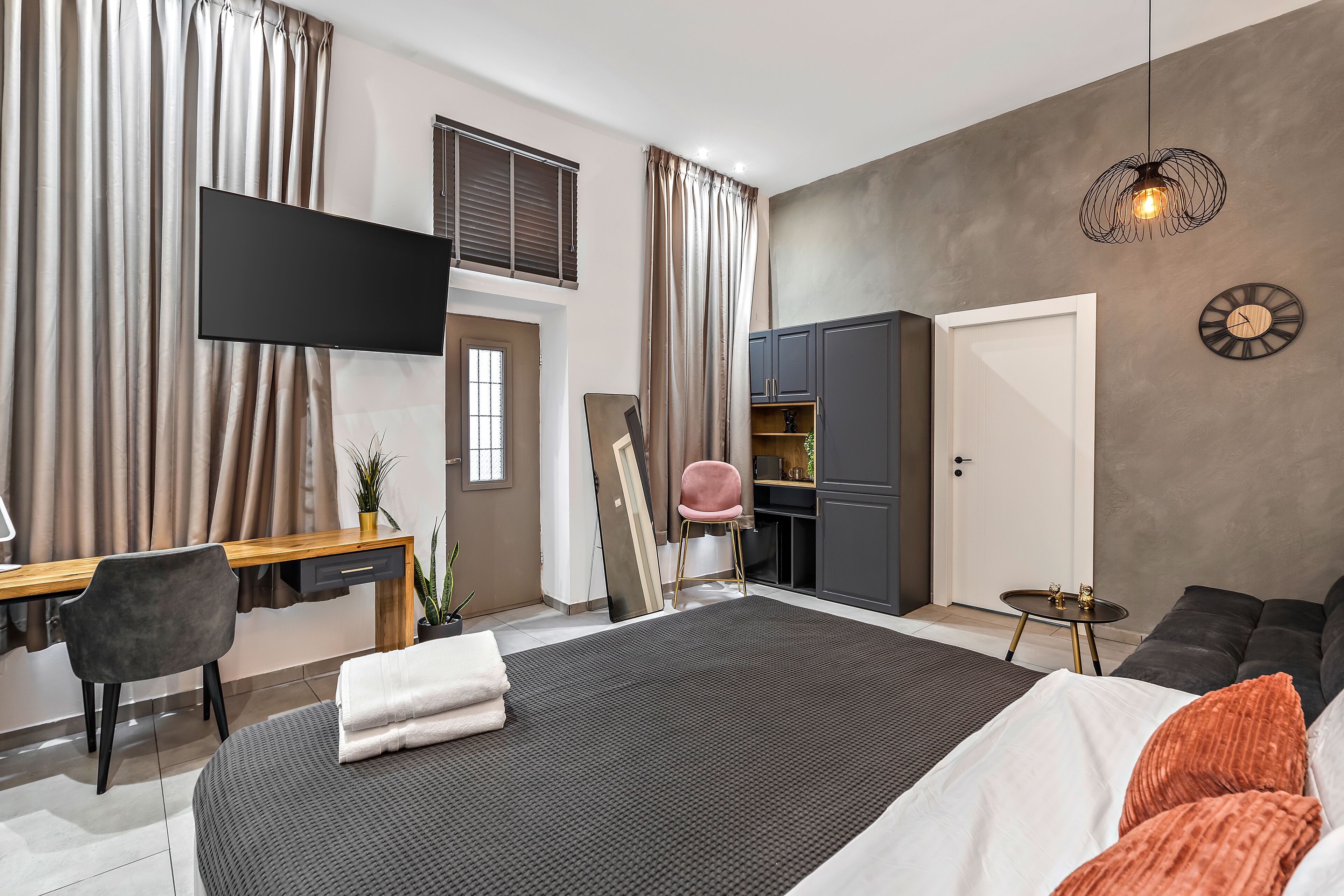 Una habitación de hotel moderna con una cama grande, un escritorio, una silla, un televisor, un armario y una mesita con objetos decorativos.