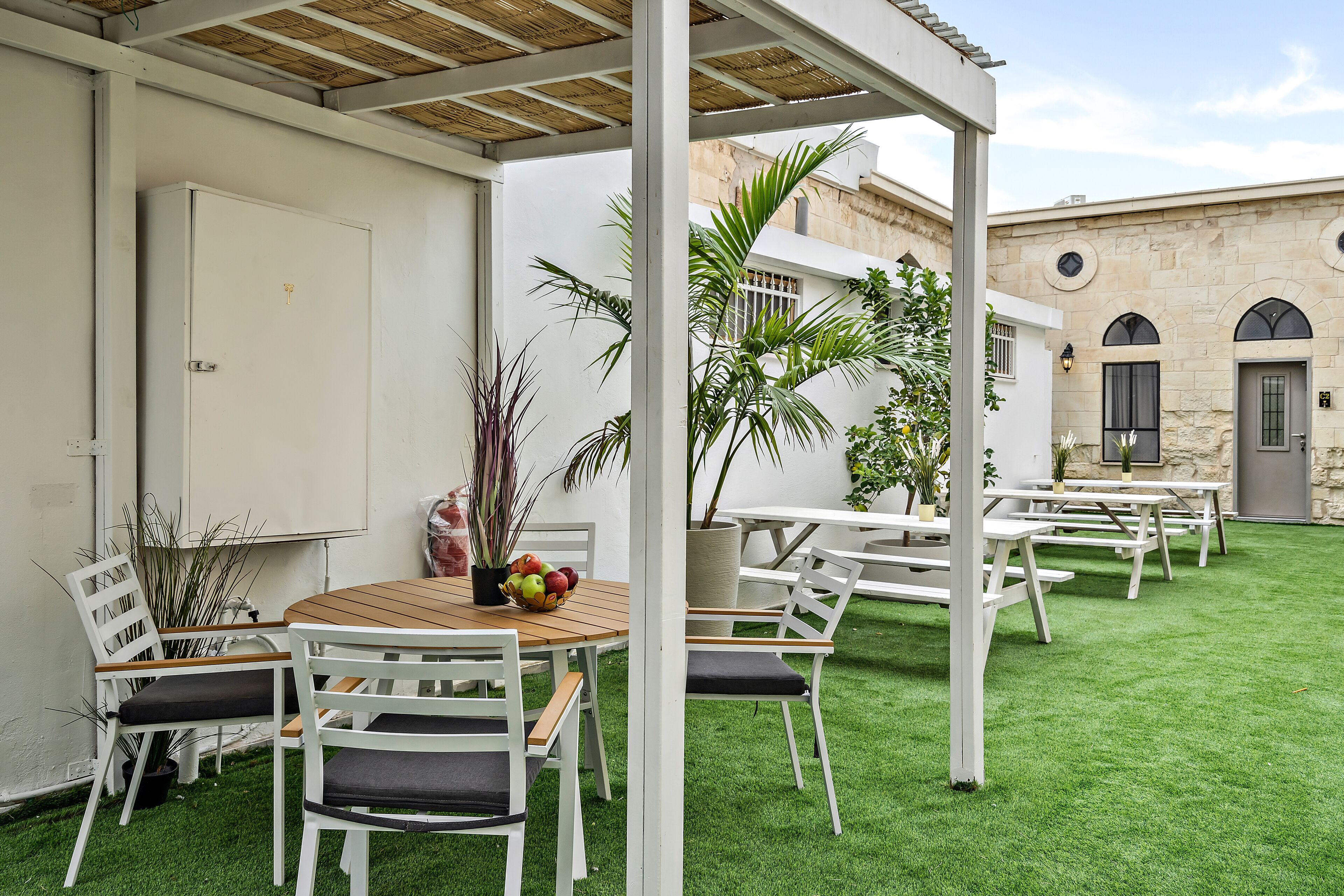 Een buitenterras met witte tafels en stoelen, omringd door groen kunstgras, en een stenen gebouw op de achtergrond.