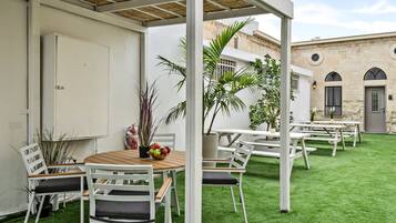 Een buitenterras met witte tafels en stoelen, omringd door groen kunstgras, en een stenen gebouw op de achtergrond.
