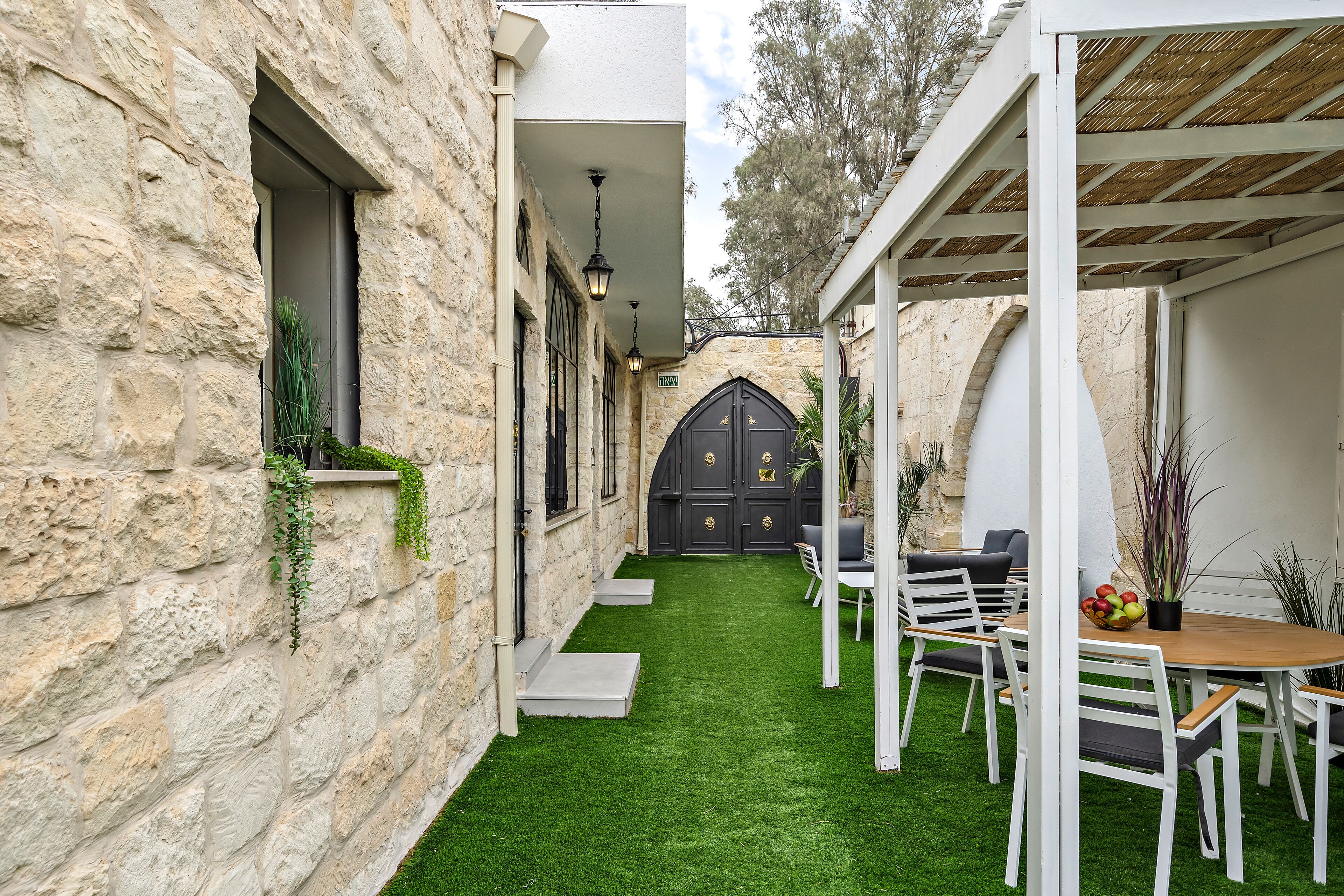 A stone building with a narrow courtyard, a wooden table with chairs, and a black door with gold accents.