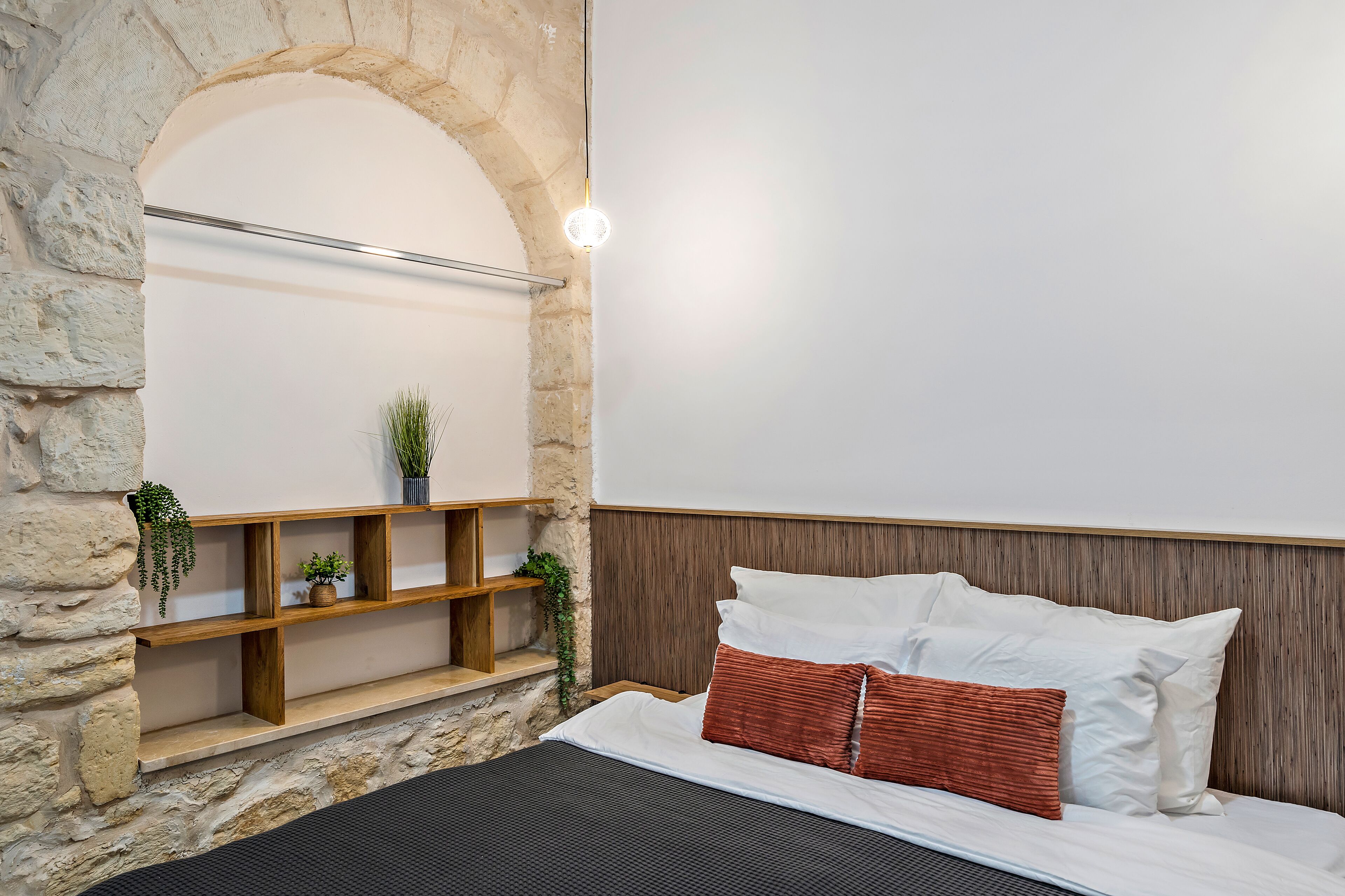 Un dormitorio con pared de piedra, estantes de madera, cama con almohadas blancas y marrones, y una lámpara colgante.