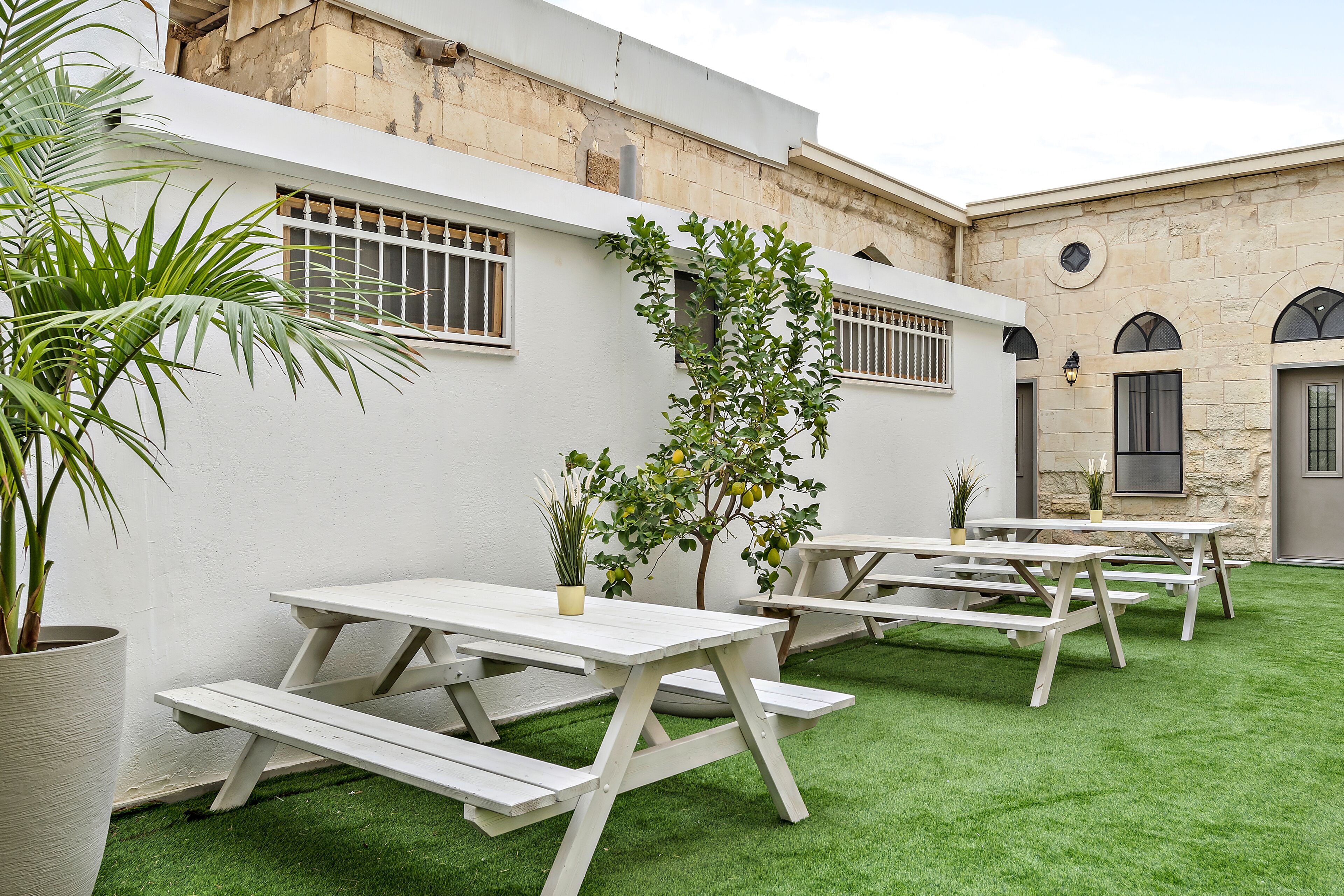 An outdoor patio area with white picnic tables and artificial grass, surrounded by stone walls and potted plants.