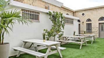 An outdoor patio area with white picnic tables and artificial grass, surrounded by stone walls and potted plants.