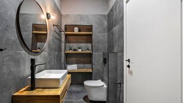 Un baño moderno con un lavabo rectangular, un inodoro y un mueble de tocador de madera. Hay un espejo sobre el lavabo y una repisa con objetos decorativos.