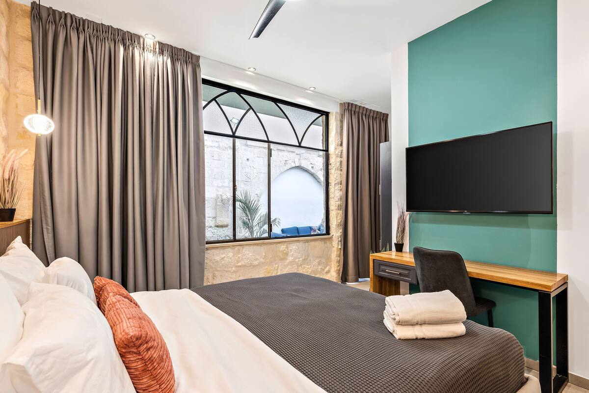 Ein Hotelzimmer mit Bett, Schreibtisch, Sessel, Fernseher und einem fenster mit Vorhängen.