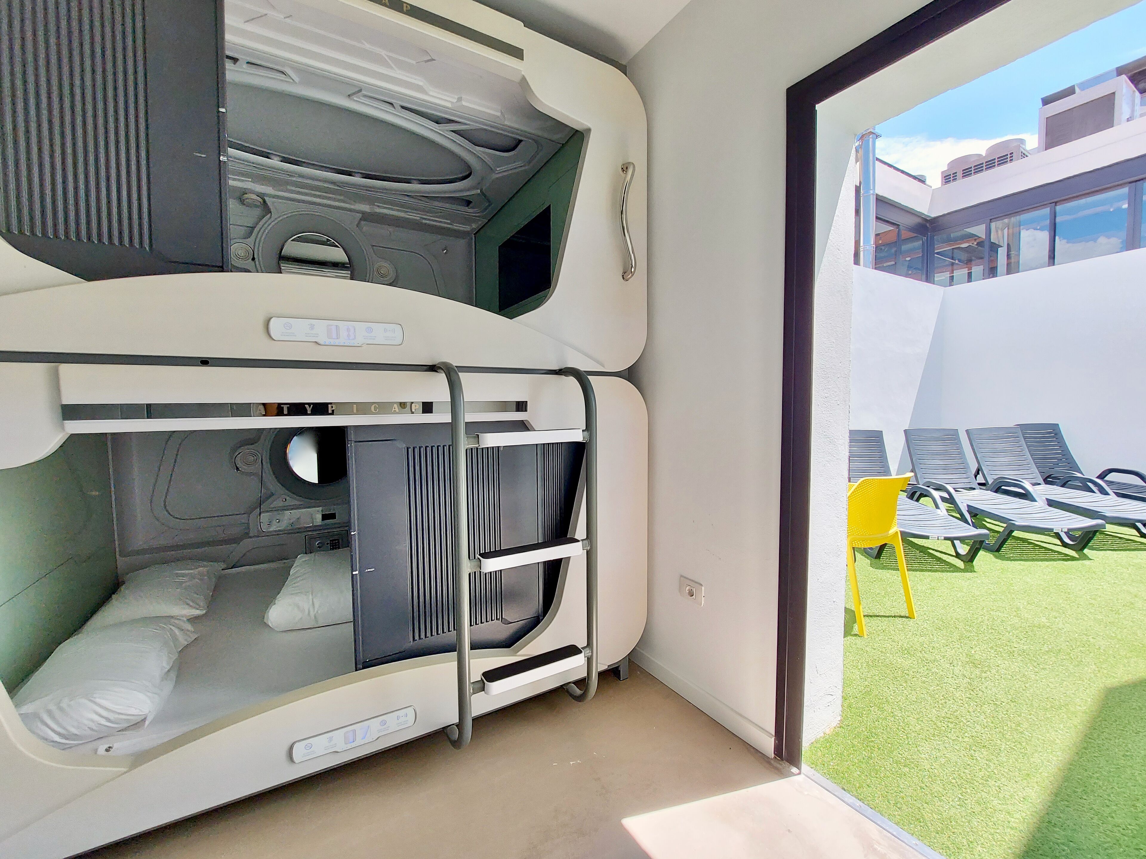A bunk bed room with a window view of a building and a grassy area with sun loungers.