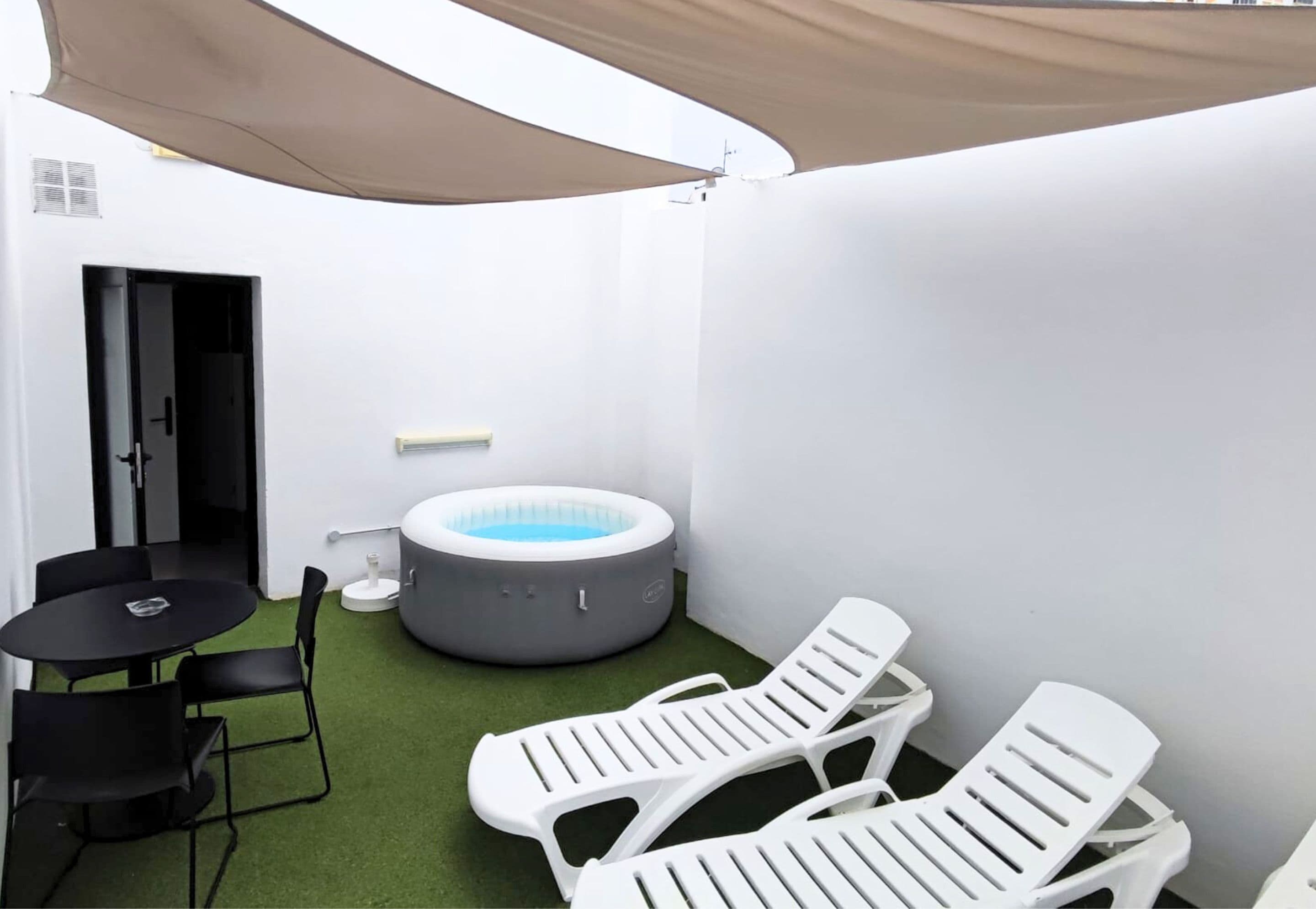 A small, enclosed outdoor area with a round hot tub, a table with chairs, and two lounge chairs.