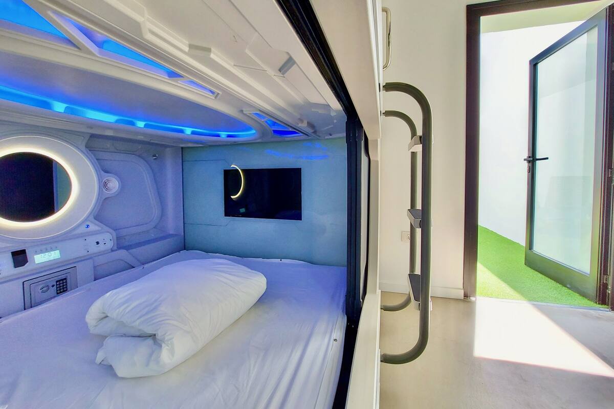 A modern room with a bed, a control panel, a small screen, and a door leading to a green outdoor area.